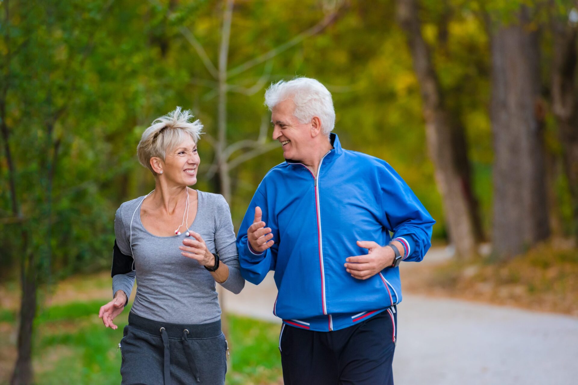 An elderly man and woman smile at each other while they're out for a jog at a park