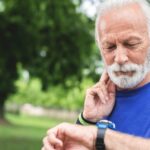 An older man in athletic clothing looks at his watch while he takes his pulse at his neck