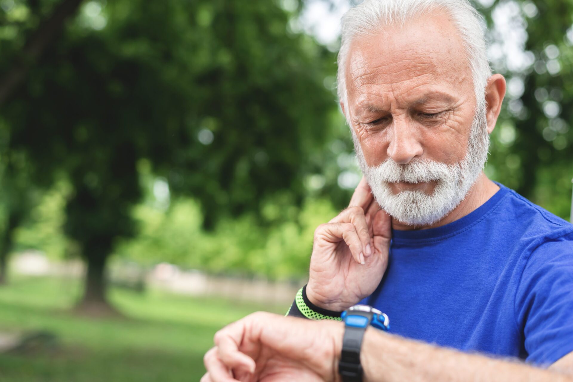 An older man in athletic clothing looks at his watch while he takes his pulse at his neck