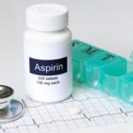An aspirin bottle sits between a weekly pill organizer and a stethoscope, atop a heart monitor chart