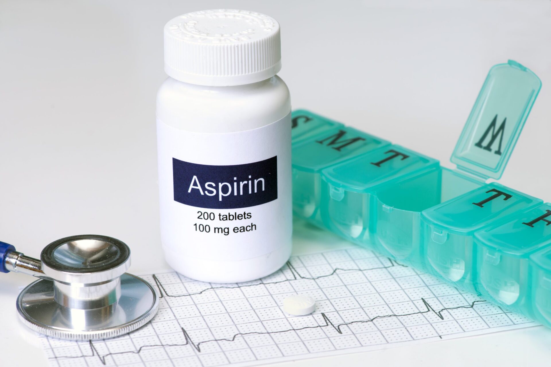 An aspirin bottle sits between a weekly pill organizer and a stethoscope, atop a heart monitor chart