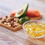 Close-up of clear yellow pills in a bowl in front of an avocado, carrot, and nuts on a cutting board