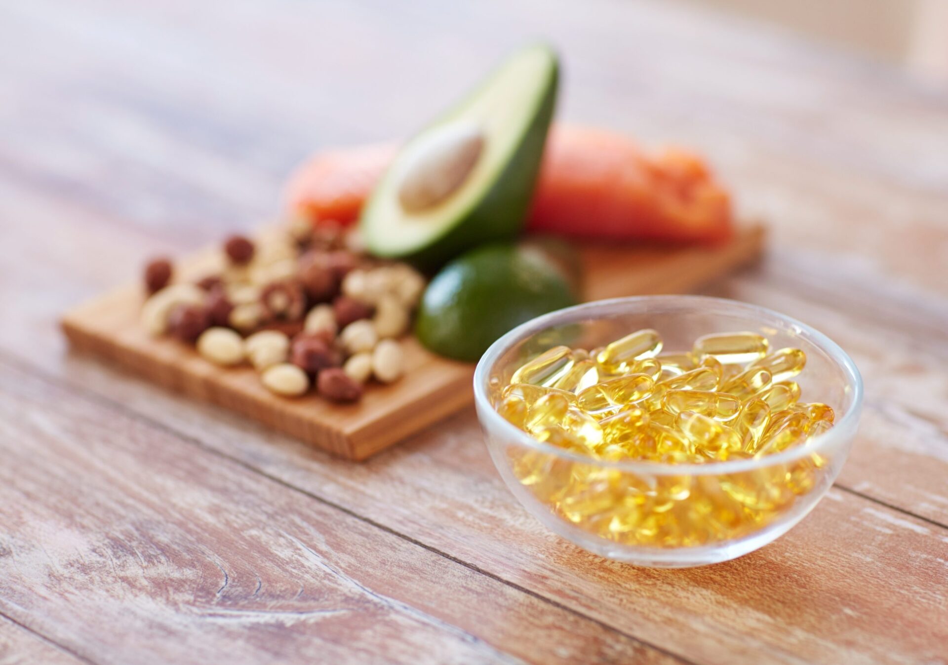 Close-up of clear yellow pills in a bowl in front of an avocado, carrot, and nuts on a cutting board
