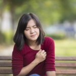 A young woman sitting on a park bench looks pained as she clenches a fist over her heart