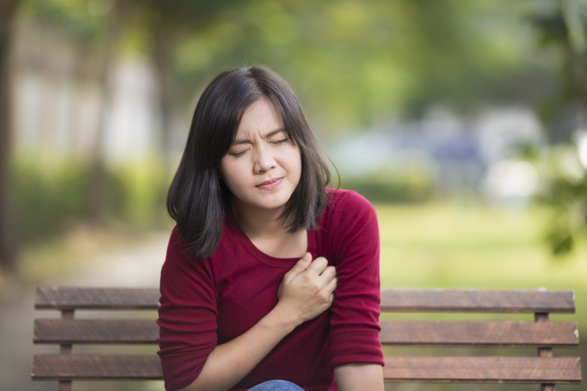 A young woman sitting on a park bench looks pained as she clenches a fist over her heart