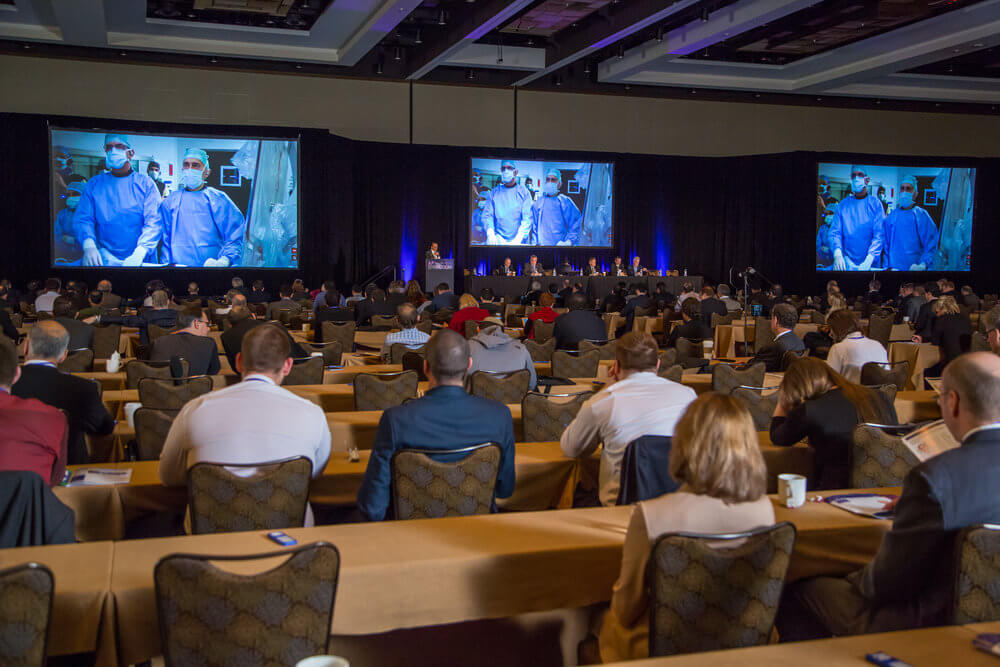 A large room filled with people at tables watching a recorded presentation and a panel of presenters