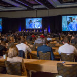 A large room full of people at tables watch a recorded presentation and a panel of presenters