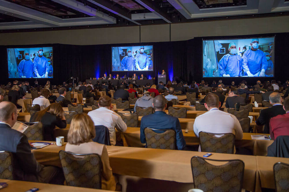 A large room full of people at tables watch a recorded presentation and a panel of presenters