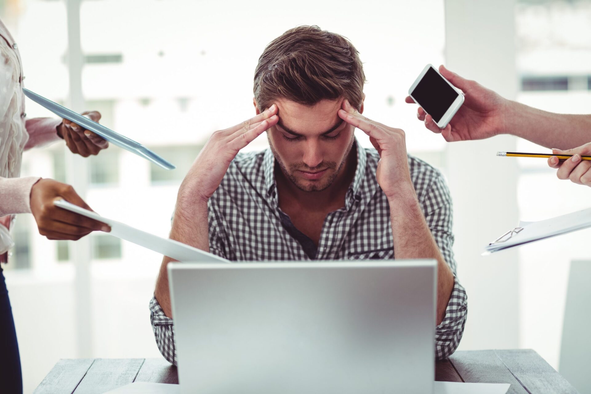 A man sits in front of a laptop looking stressed while a hand holds a cell phone out to him