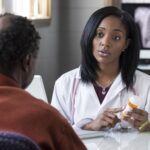 A female doctor with dark brown skin sits behind a desk, explaining a medication to a male patient