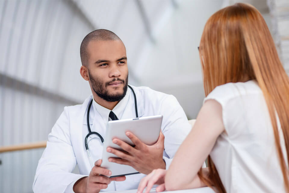A male doctor with light brown skin talking to a female patient while holding a tablet